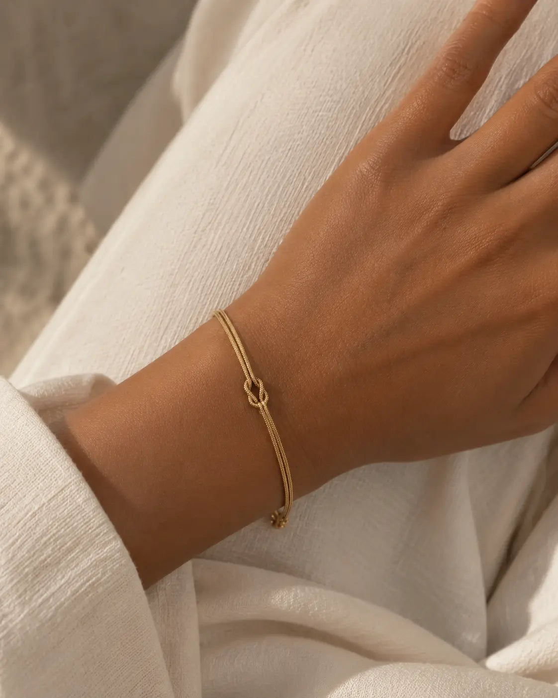 Gold-tone double-chain bracelet with a central knot, close-up on a wrist resting on light fabric.