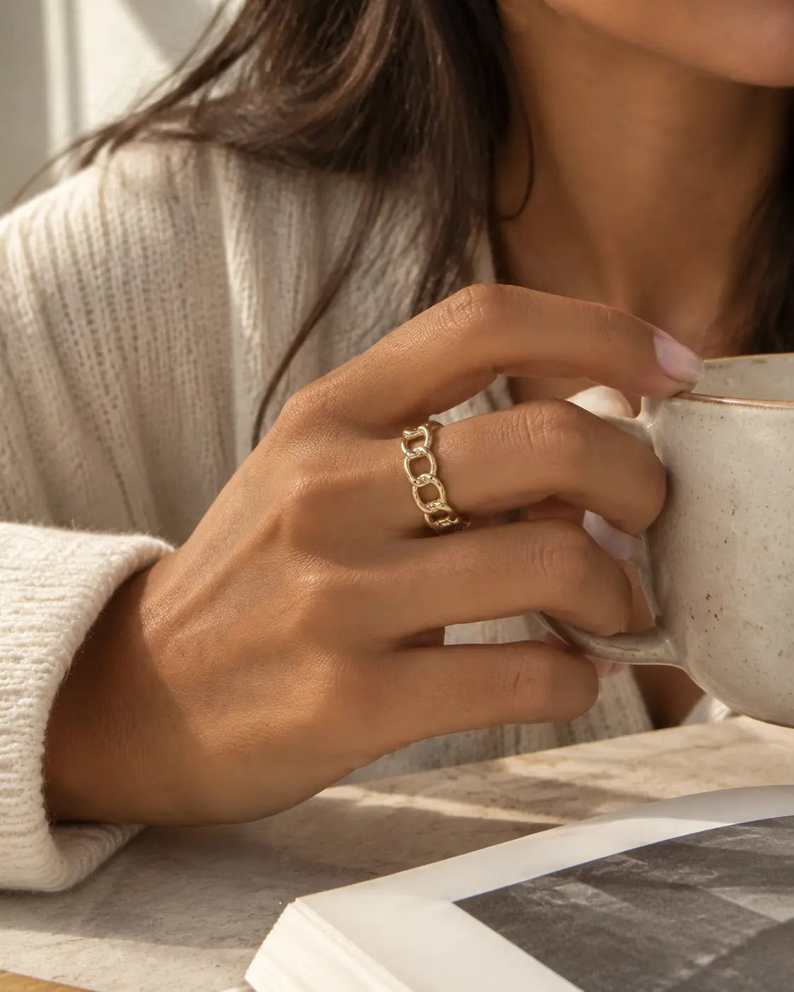Gold-tone open ring with oval chain links, worn on a hand holding a cup.