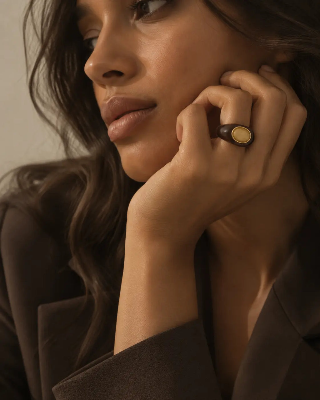 Brown ring with a pale oval inset and gold-tone rim, worn with a hand against the cheek.