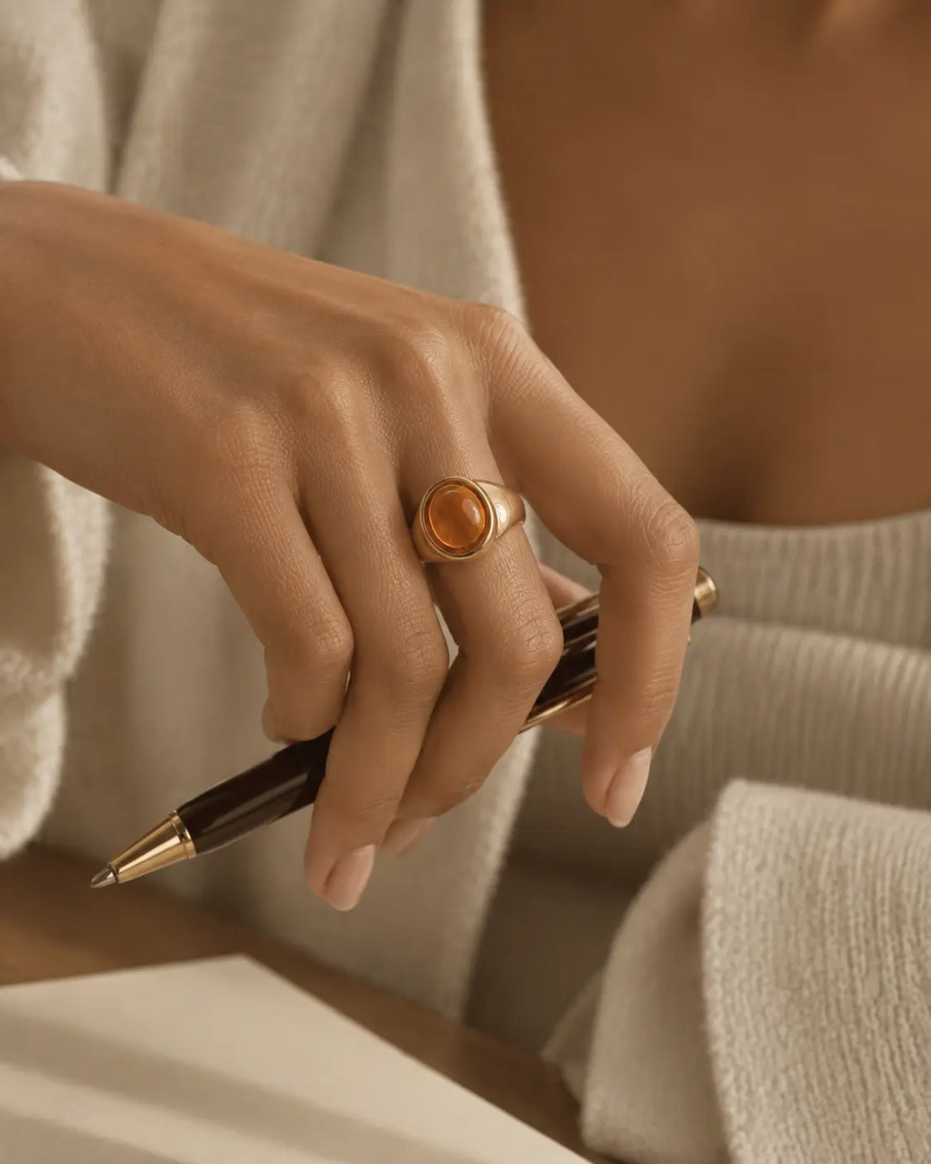 Gold-tone signet ring with a raised warm-brown oval accent, worn on a hand holding a pen over a book.