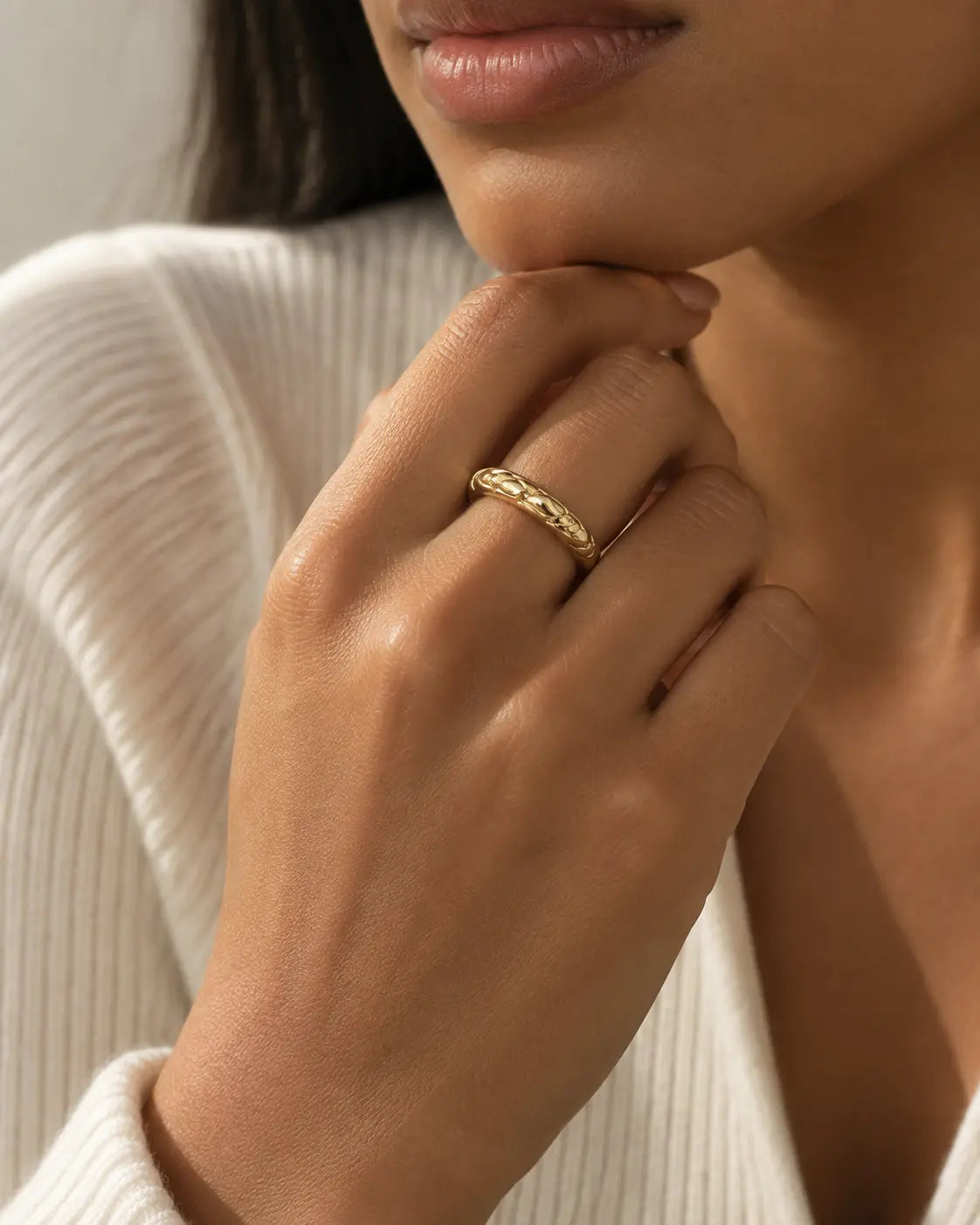 Gold-tone band ring with interwoven rounded twists, worn on a hand near the chin beside cream knitwear.