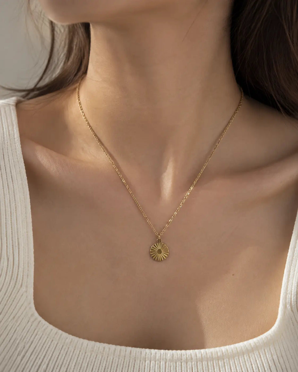 Gold-tone necklace with a round sunburst coin pendant, close-up worn with a white square-neck top.