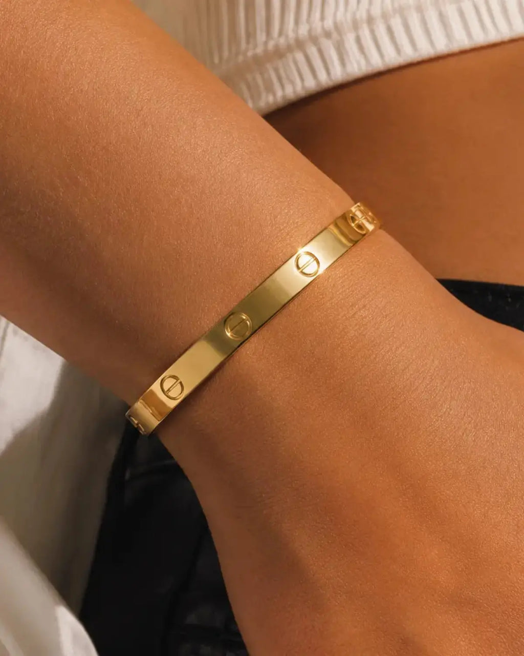 Plain gold-tone bangle with engraved screw motifs, close-up on the wrist beside a white cuff.