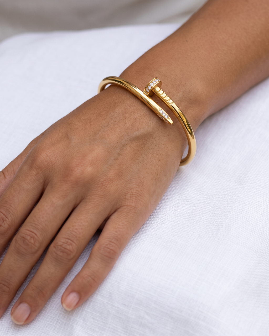 Gold-tone nail-shaped bangle with stone accents, worn against white fabric with the nail head and tip visible.