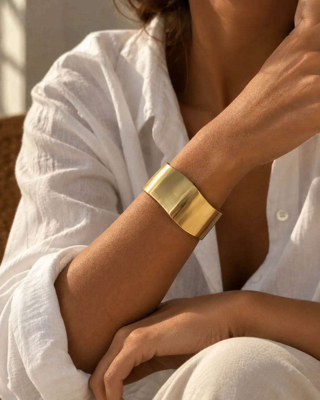 Gold-tone wide bangle with a satin-looking surface and wavy edges, worn on a raised arm in a seated portrait.