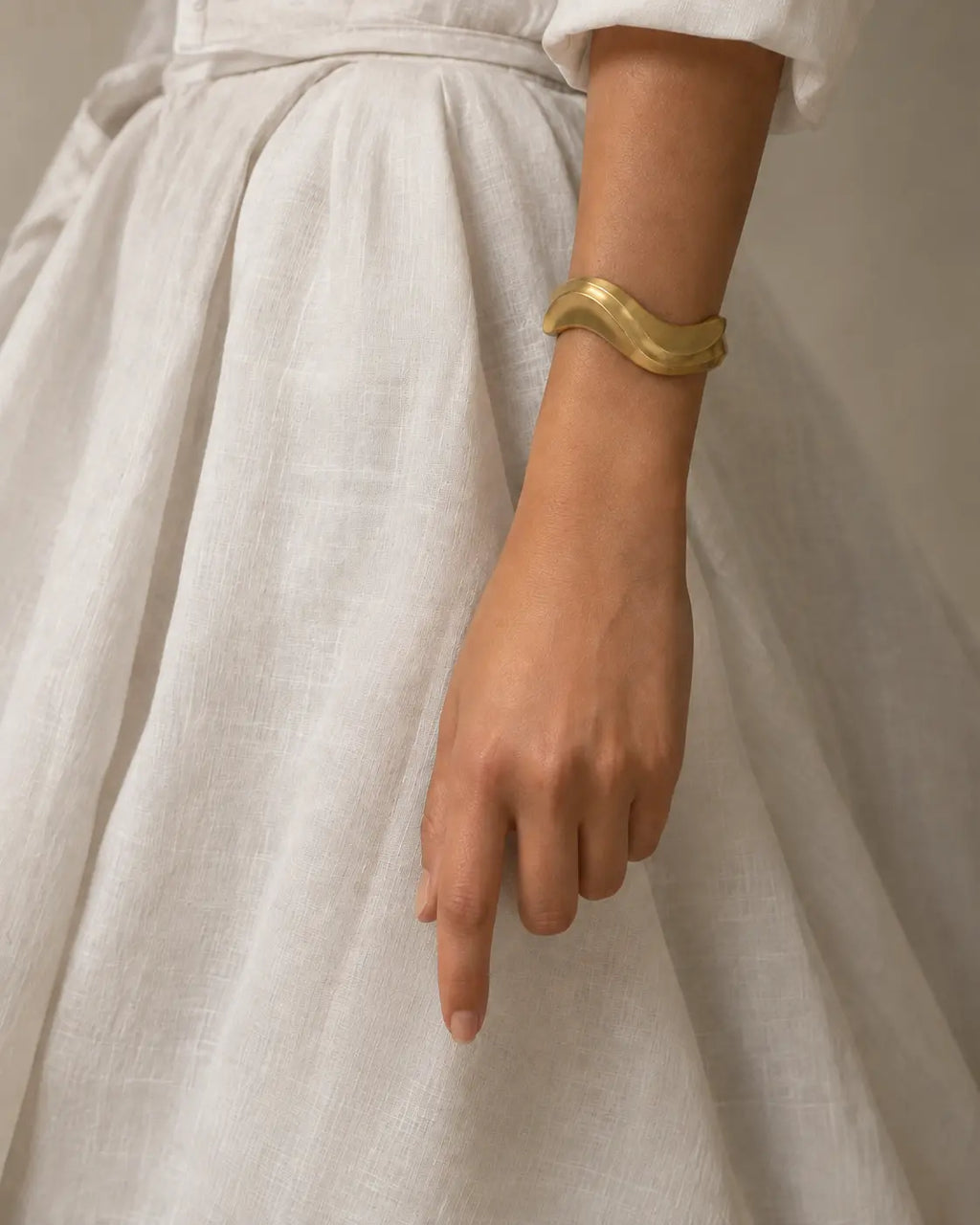 Gold-tone sculptural bangle with an undulating outline, worn beside the tied waist of a white dress.