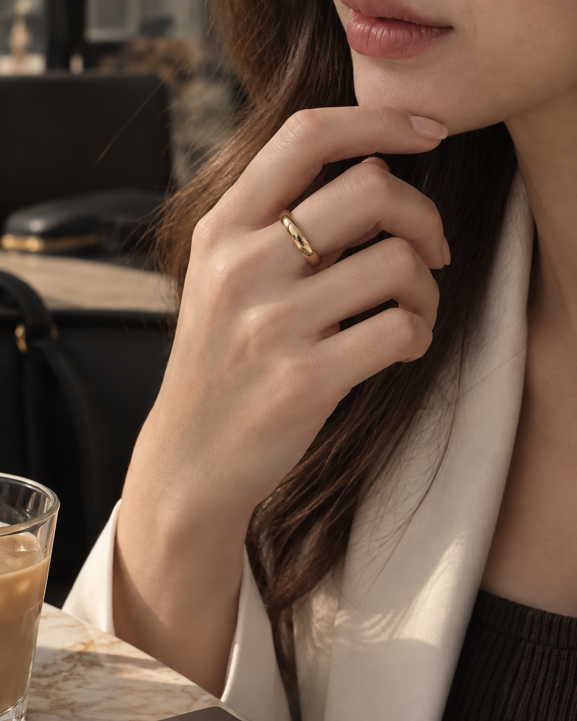 Gold-tone smooth dome band ring, worn on a hand near the face beside a white jacket.