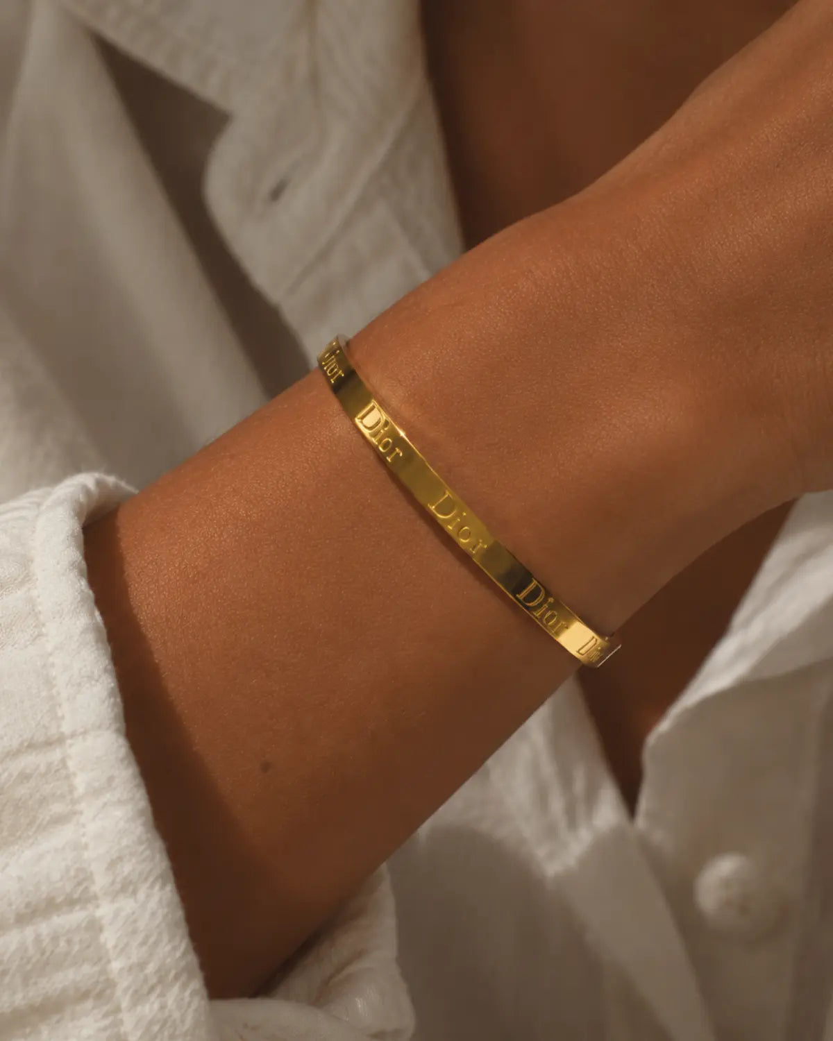 Gold-tone bangle with engraved lettering, worn at the wrist beside an open white shirt.