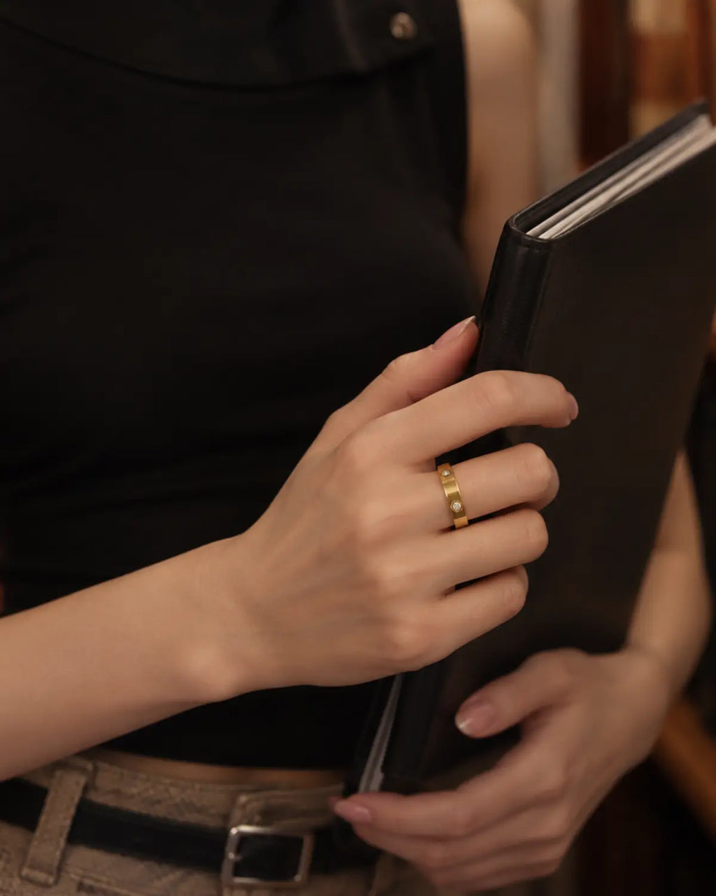 Gold-tone band ring with spaced round bezel-set stone accents, worn on a hand holding a black clutch.