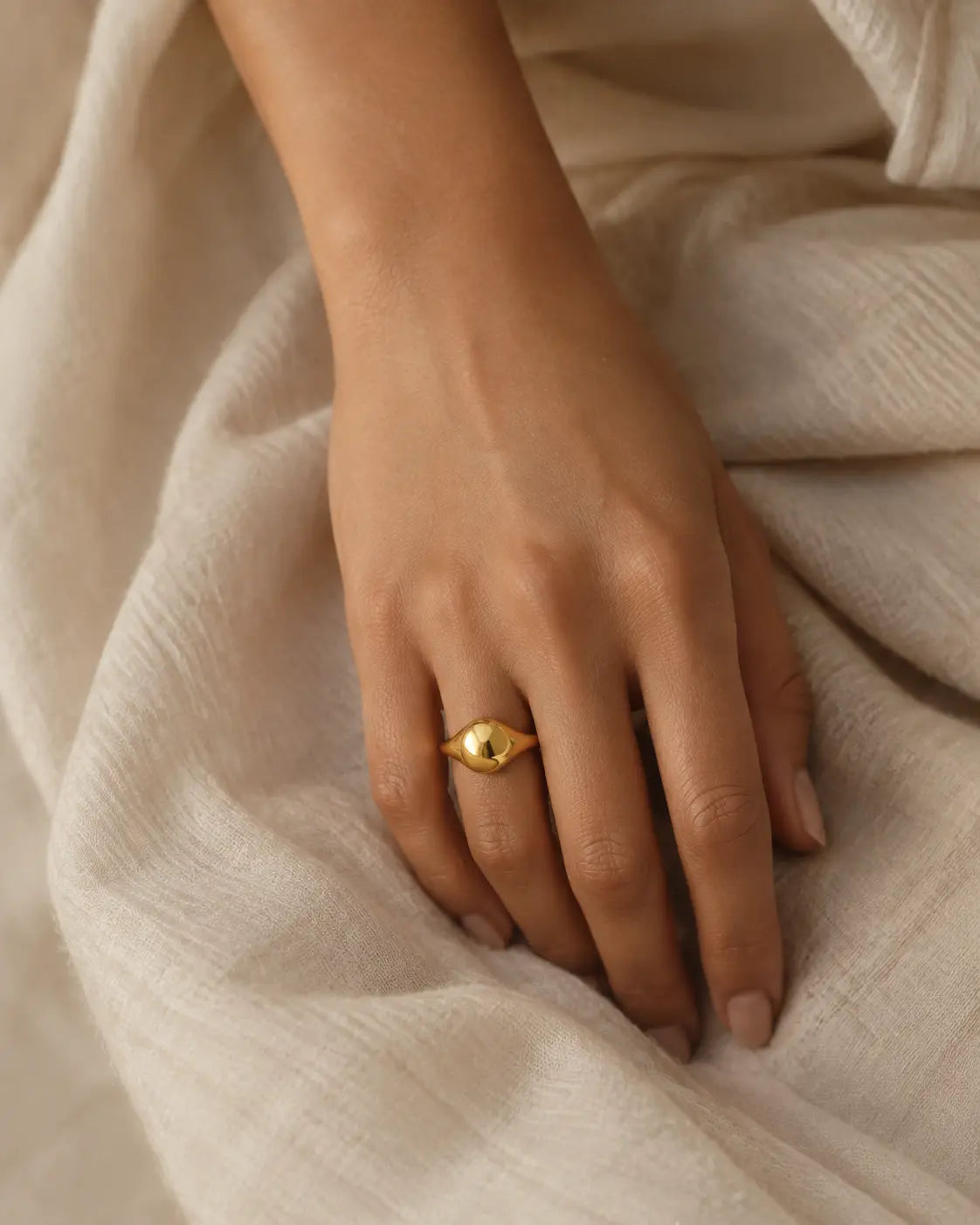 Gold-tone ring with an asymmetric domed front, worn on a hand resting on light fabric.