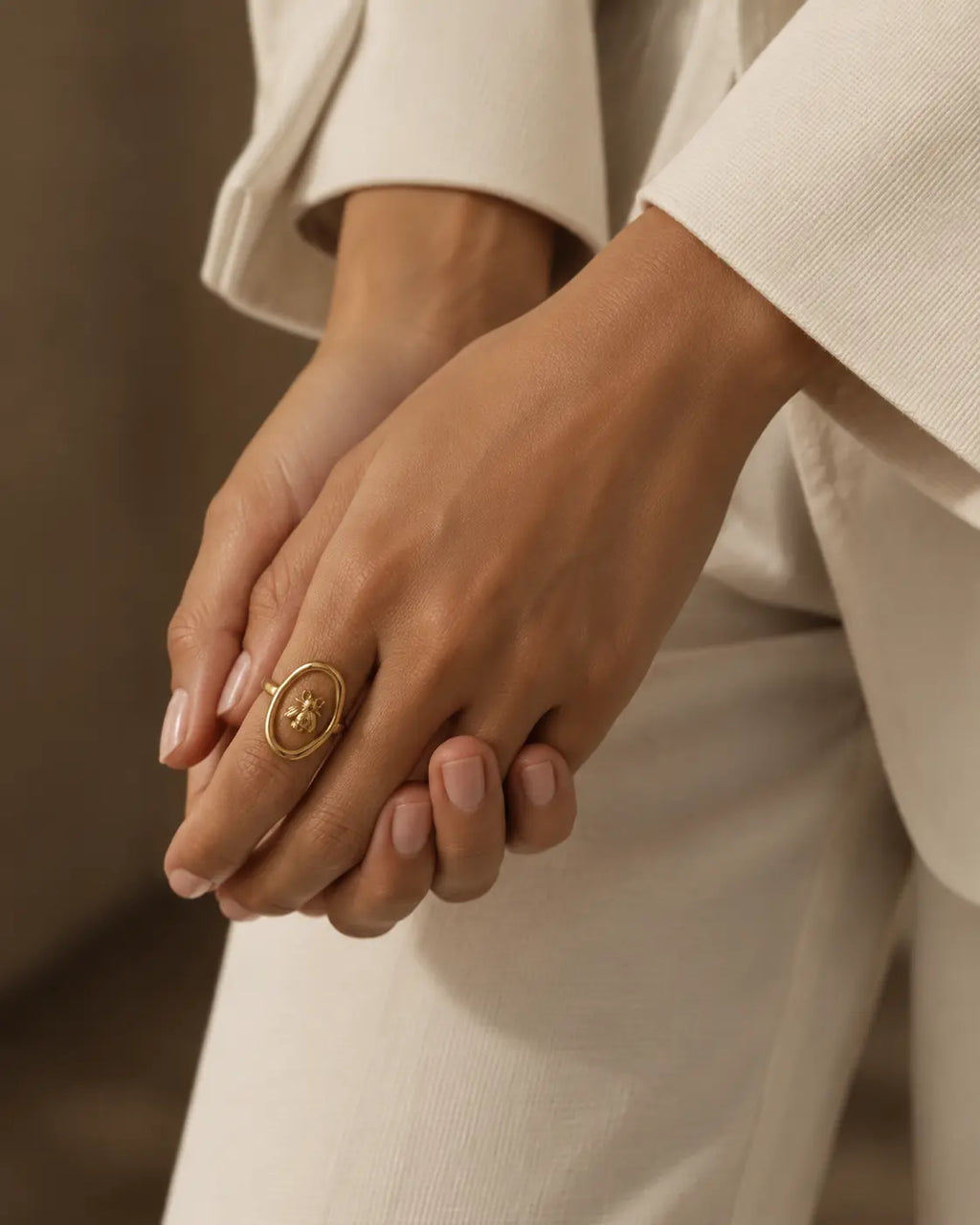 Gold-tone ring with a bee inside an open oval frame, worn with hands resting together beside white sleeves.