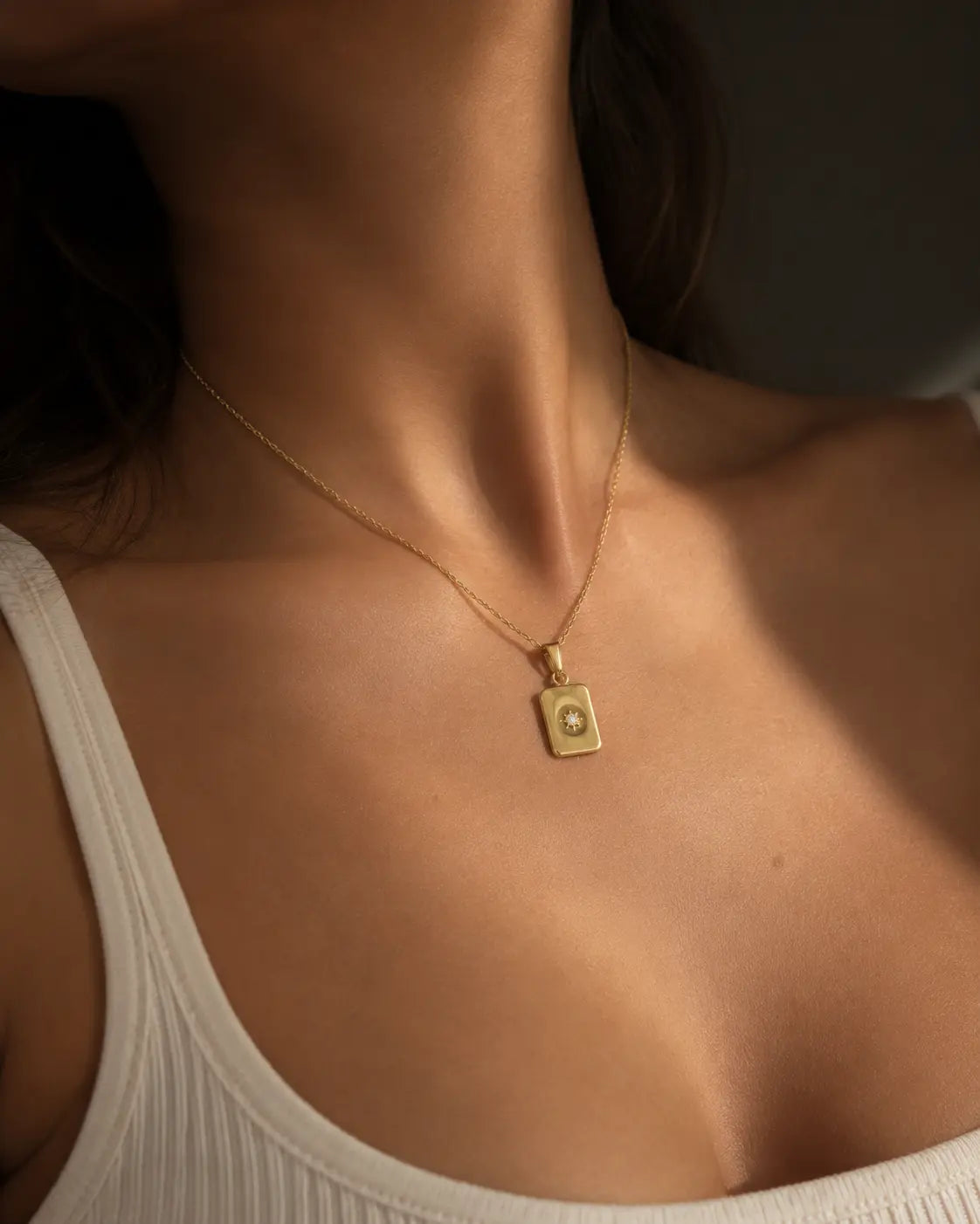 Gold-tone necklace with a rectangular celestial-motif tag pendant, worn with a white square-neck top.