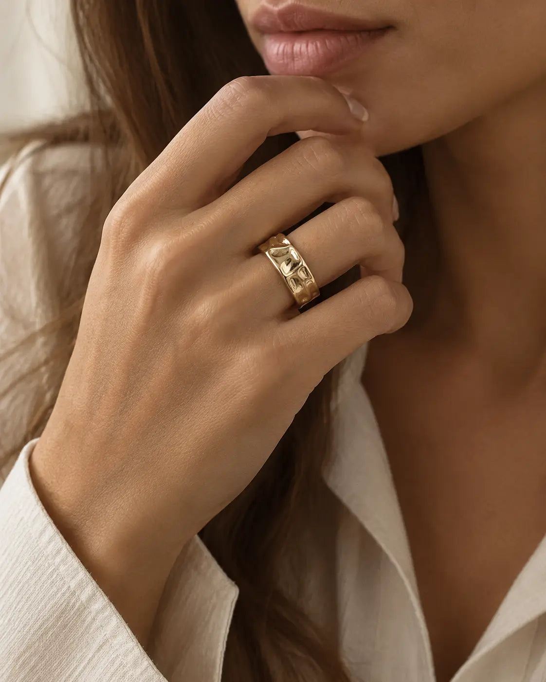 Gold-tone open band ring with a folded surface, worn on a hand near the chin.