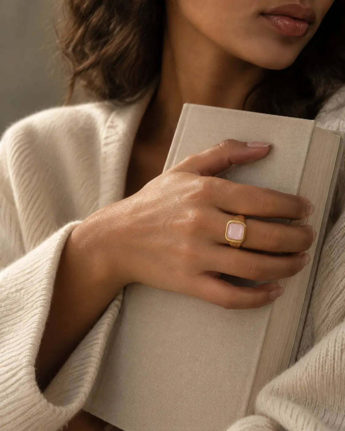 Gold-tone ring with a pale pink cushion-shaped accent, worn on a hand holding a book.