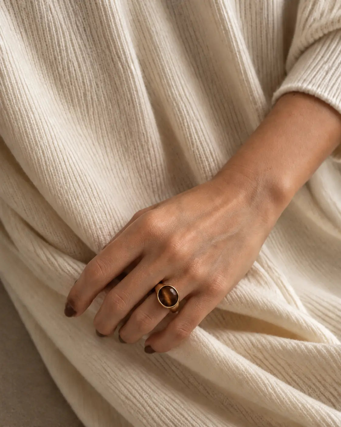 Gold-tone ring with a horizontal brown oval accent, worn on a hand resting against cream knitwear.