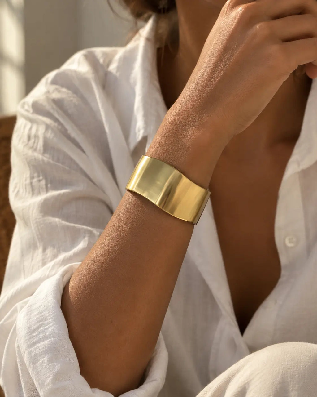 Gold-tone wide bangle with a satin-looking surface and wavy edges, worn beside a white shirt cuff.
