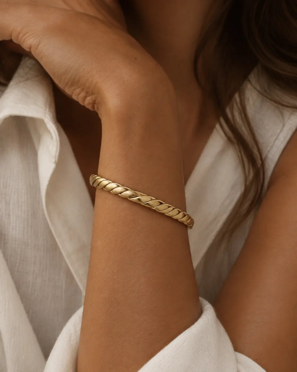 Gold-tone open bangle with a thick rope-twist pattern, close-up on a wrist near the chin.