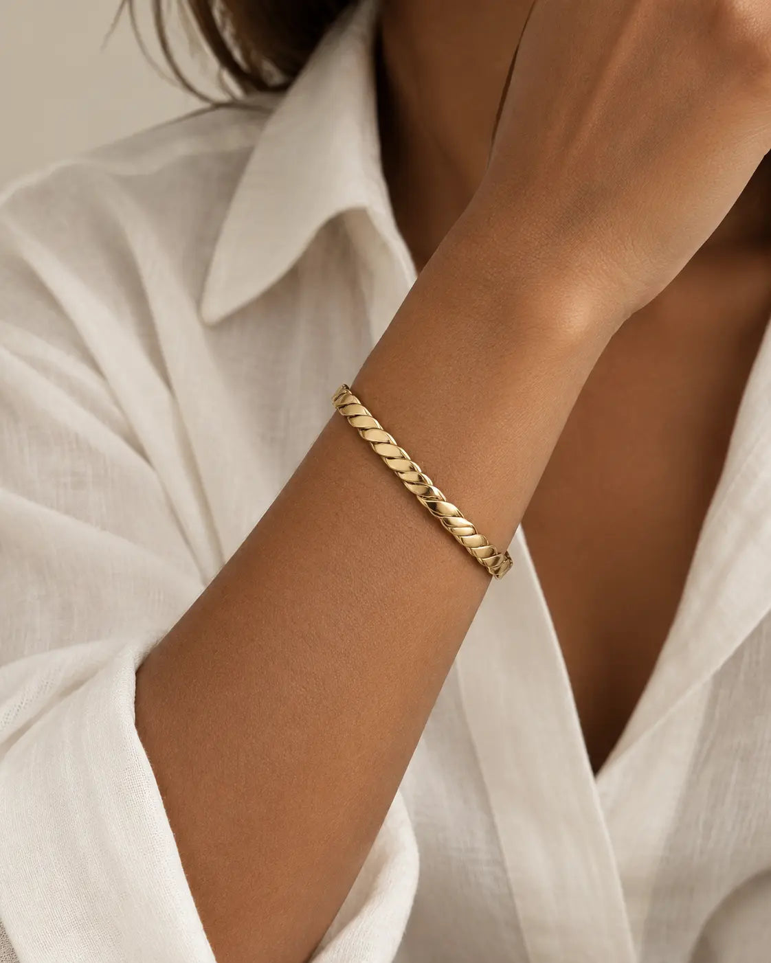 Gold-tone open bangle with a thick rope-twist pattern, worn on a raised wrist beside a white collar.