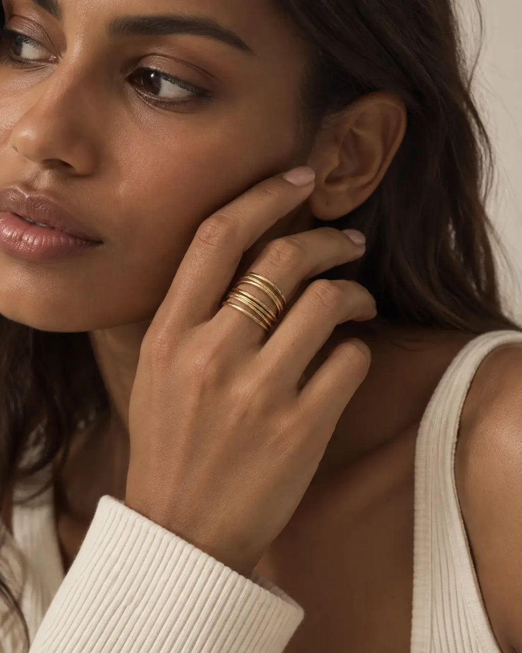 Gold-tone ring with multiple crisscrossing rails, worn on a hand near the face beside cream knitwear.