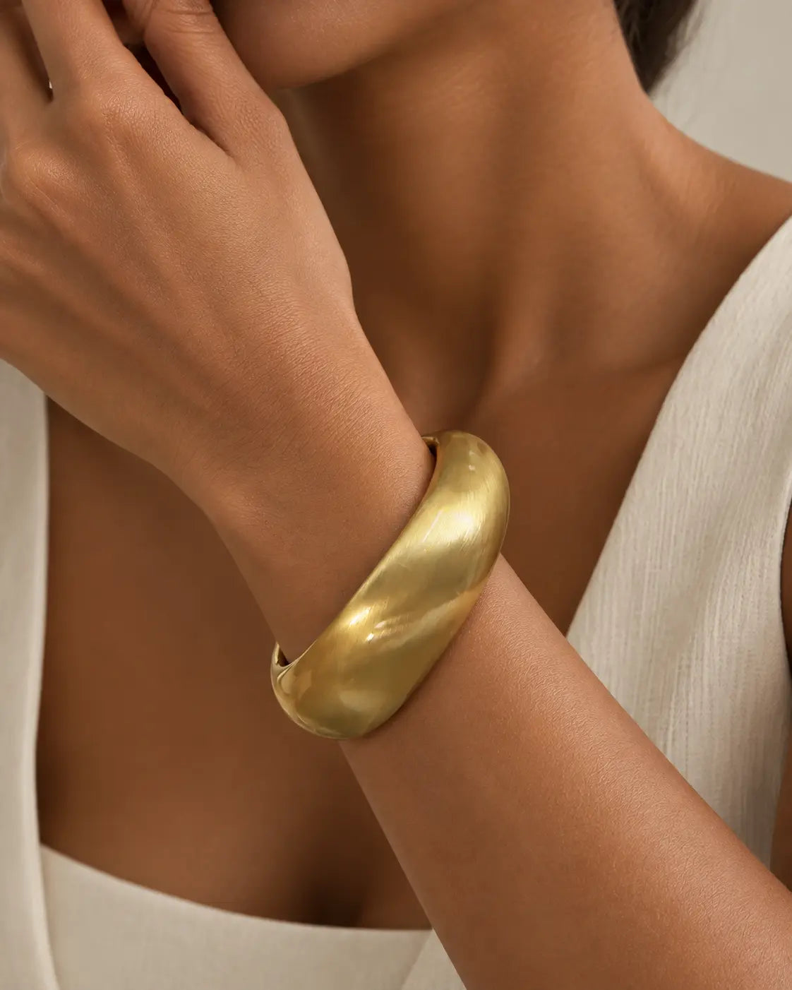 Gold-tone rounded contour bangle with a brushed-looking surface, worn on a wrist near the chin.