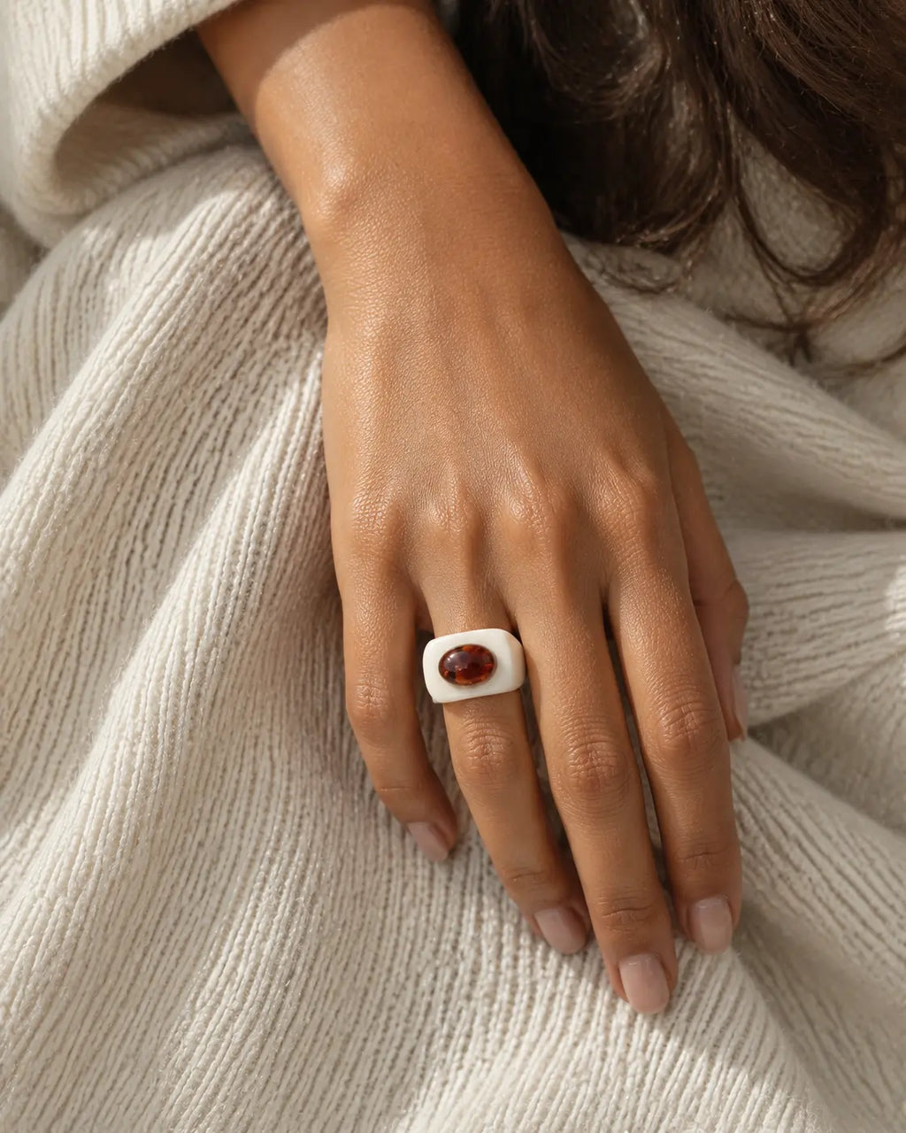 White ring with a contrasting brown oval accent, worn on a hand resting against cream knitwear.