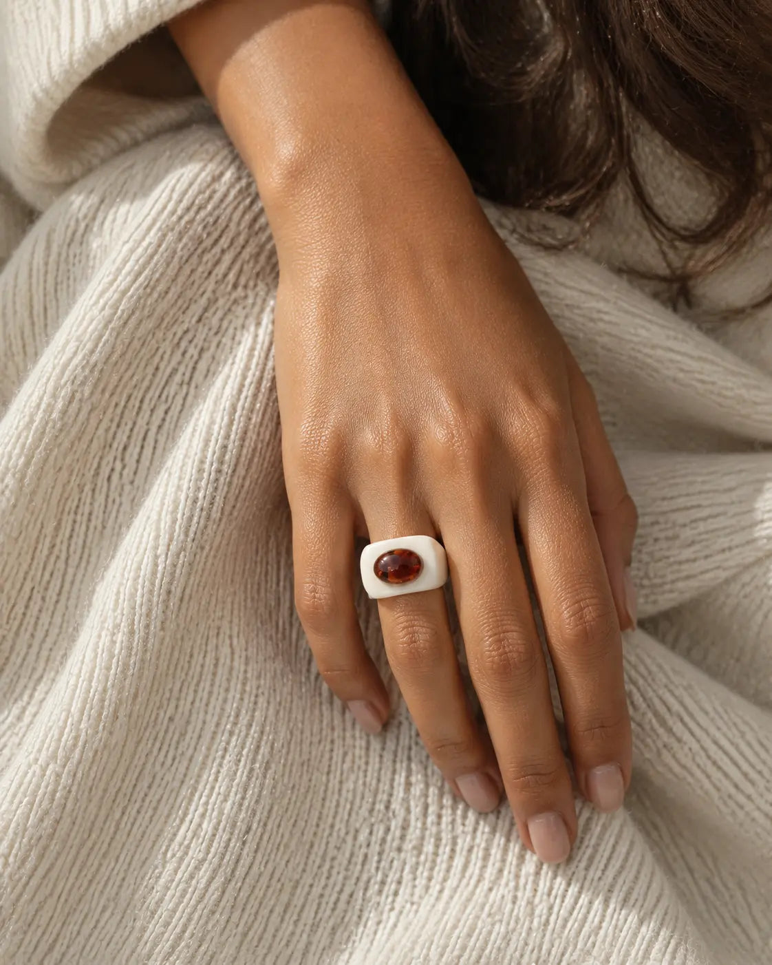White ring with a contrasting brown oval accent, worn on a hand resting against cream knitwear.