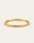 Gold-tone bangle with repeated engraved lettering, shown at an angle.