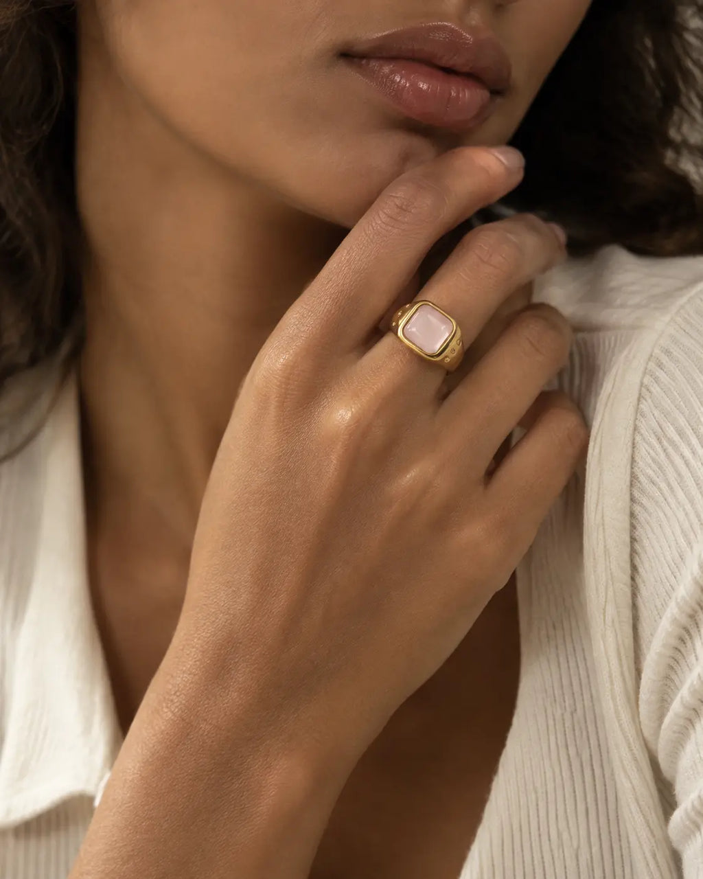 Gold-tone ring with a pale pink cushion-shaped accent, worn on a hand near the chin.