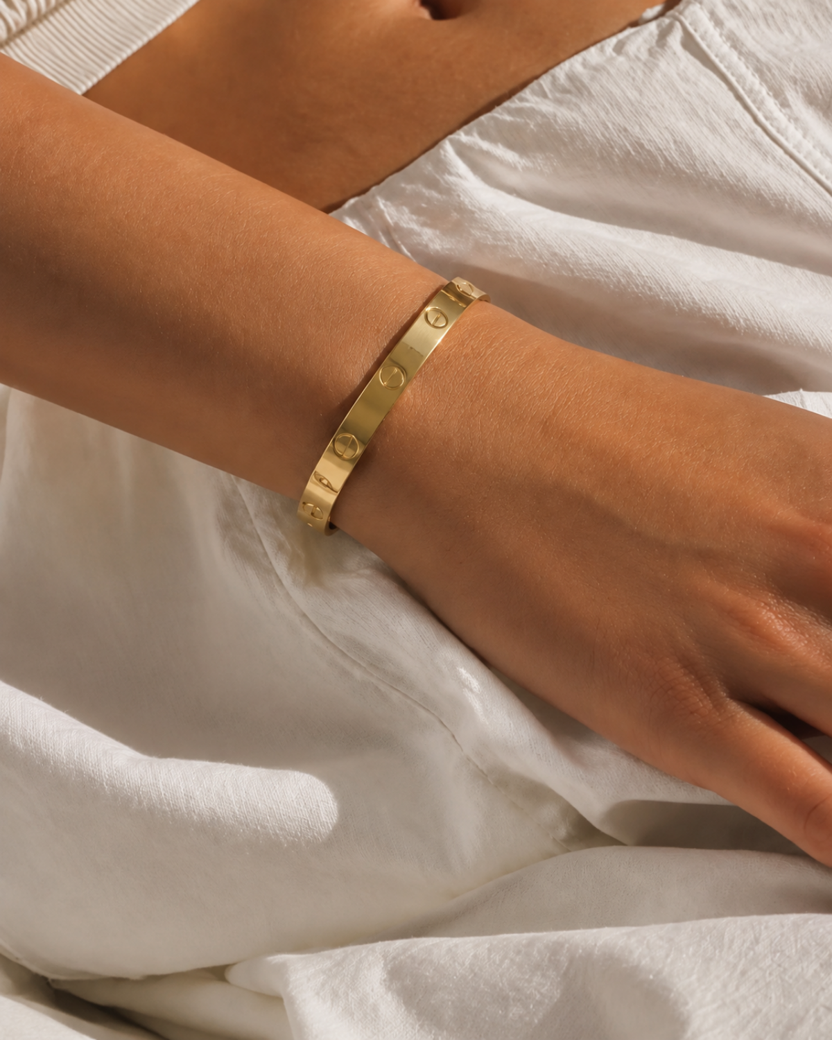 Plain gold-tone bangle with engraved screw motifs, worn on a wrist resting against white fabric.