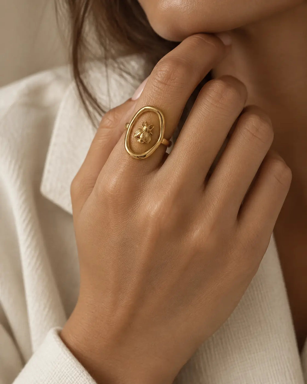 Gold-tone ring with a bee inside an open oval frame, worn on a hand near the chin.