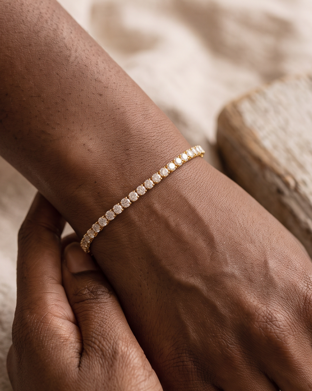 Gold-tone tennis bracelet with a continuous row of clear stone accents, worn with hands resting together.