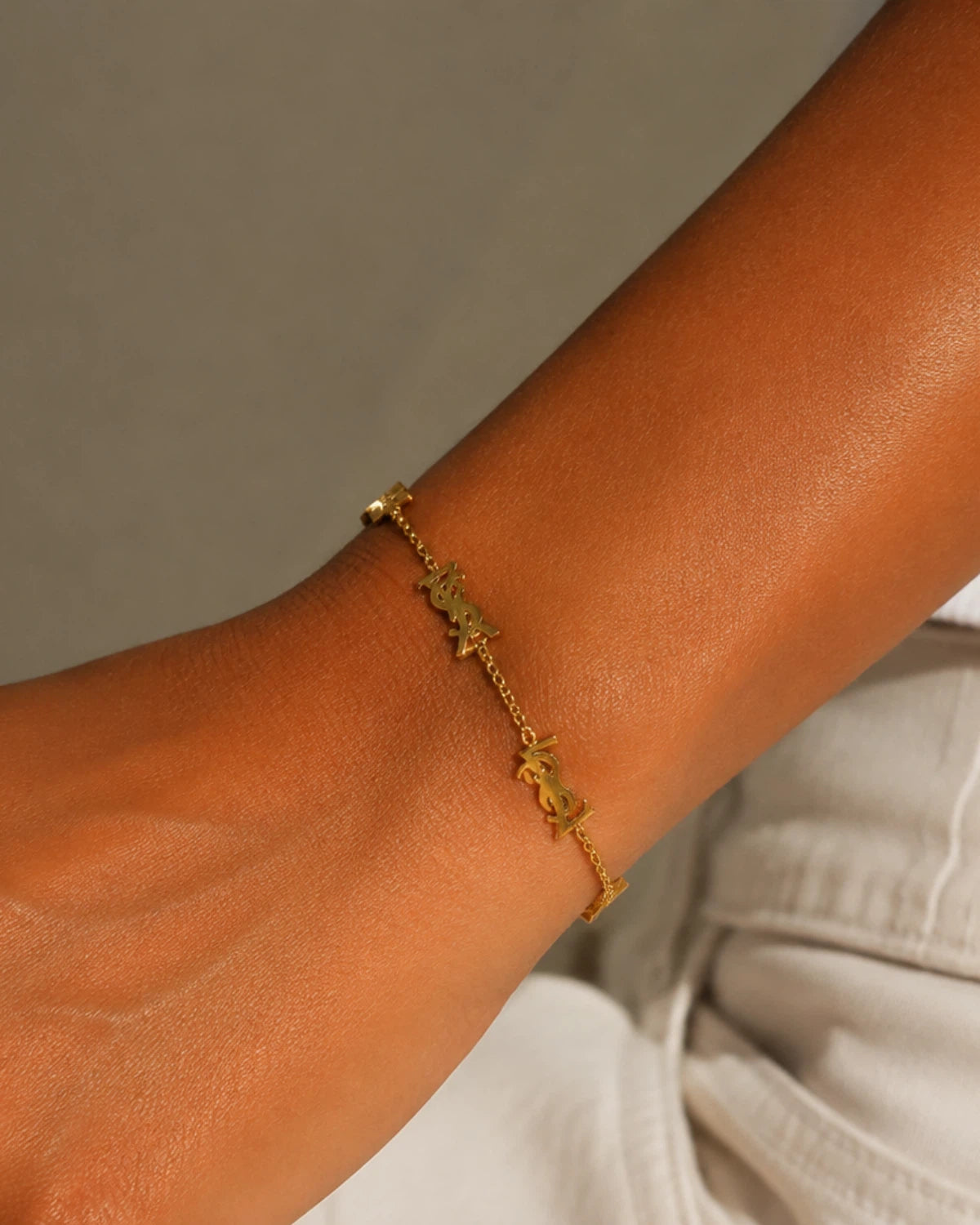 Gold-tone chain bracelet with repeated monogram-style stations, angled view on a wrist resting on light fabric.