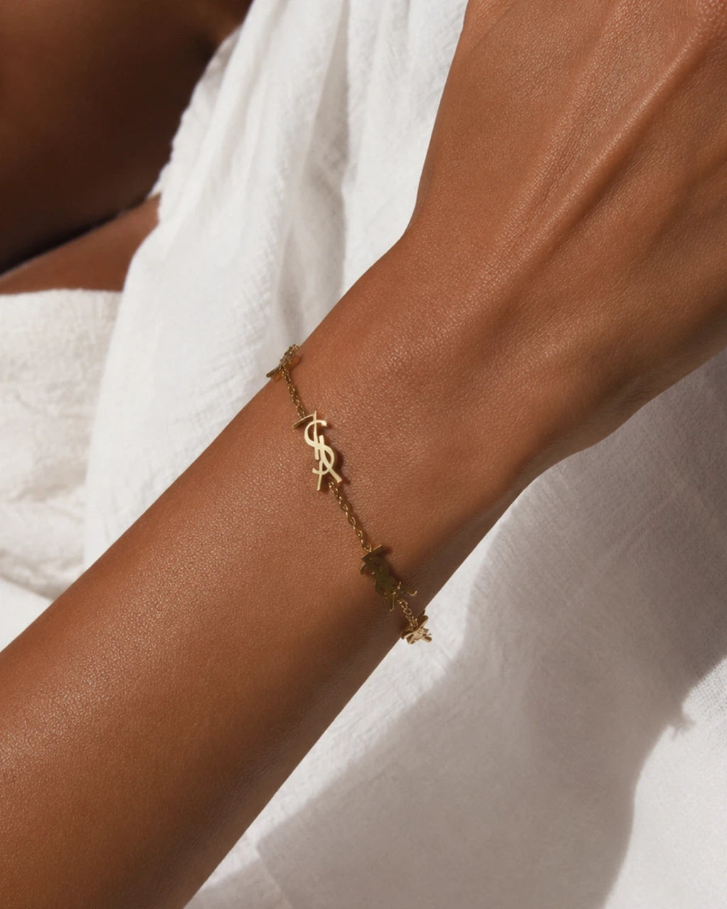 Gold-tone chain bracelet with repeated monogram-style stations, worn across the wrist against white fabric.