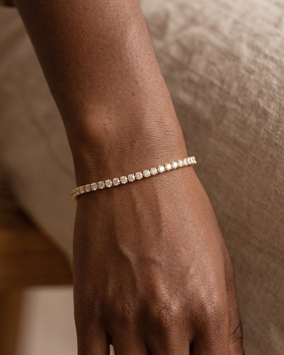 Gold-tone tennis bracelet with a continuous row of clear stone accents, worn across the wrist above the back of the hand.