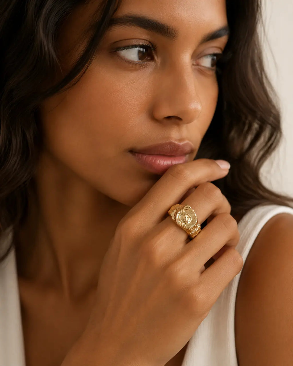 Gold-tone signet ring with an irregular molten-like texture, worn on a hand near the chin.