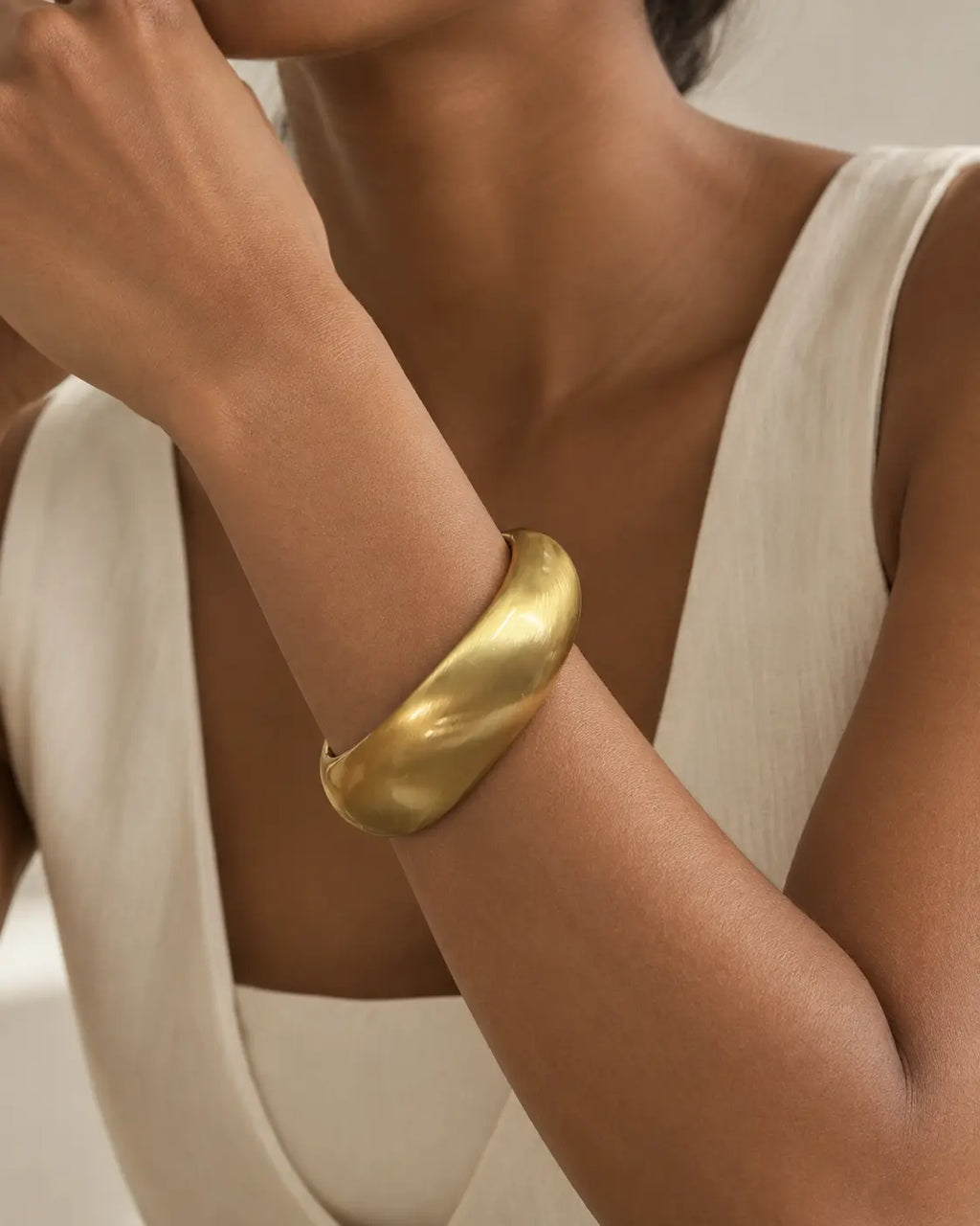 Gold-tone rounded contour bangle with a brushed-looking surface, close-up on the wrist beside white knitwear.