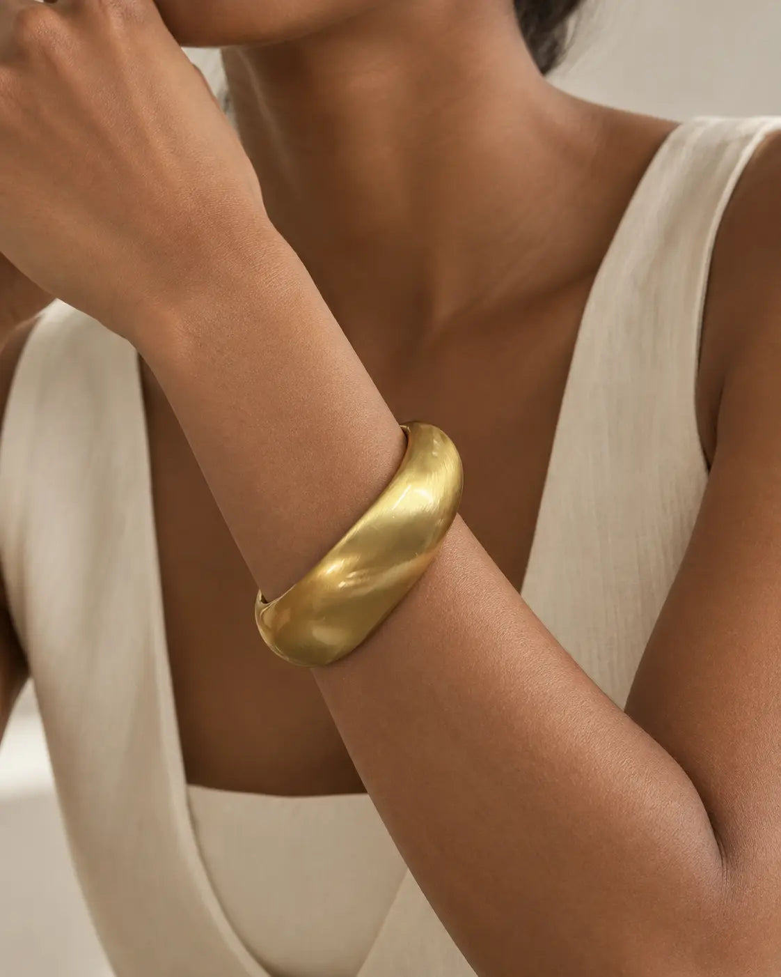 Gold-tone rounded contour bangle with a brushed-looking surface, close-up on the wrist beside white knitwear.