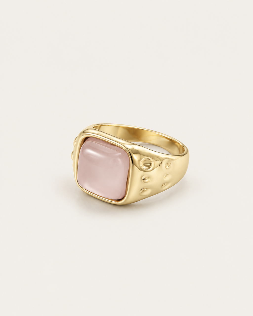 Gold-tone ring with a pale pink cushion-shaped accent, angled view showing the textured shoulders.