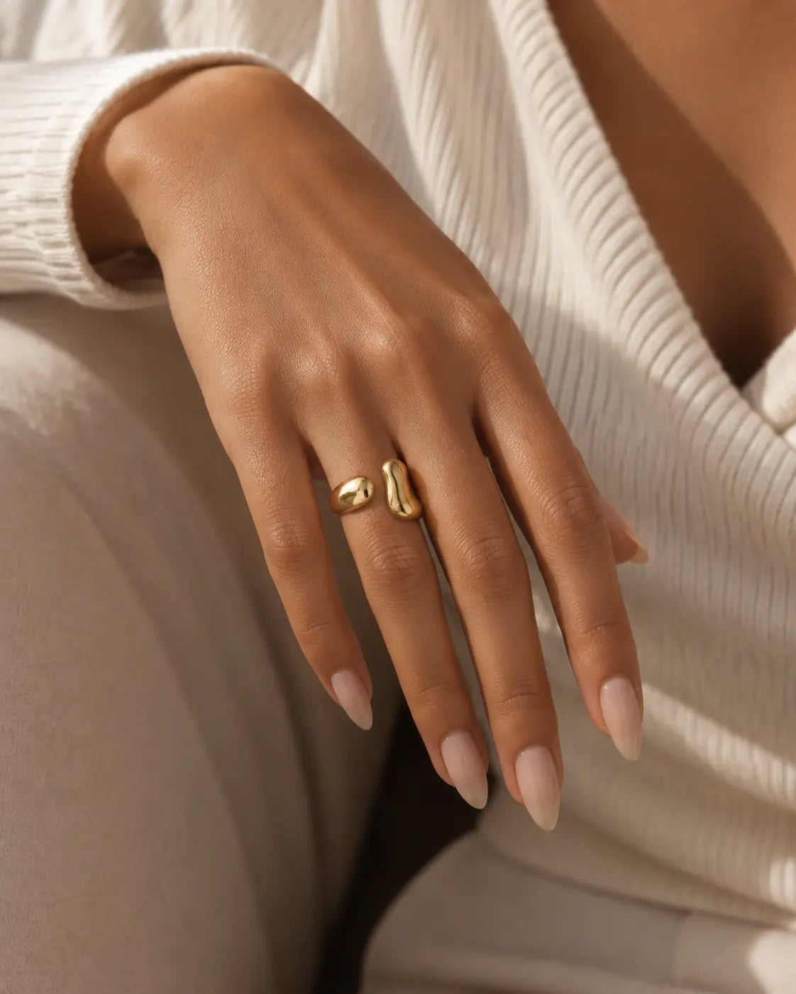 Gold-tone bypass ring with two rounded drop-shaped ends, worn on a hand resting against cream knitwear.