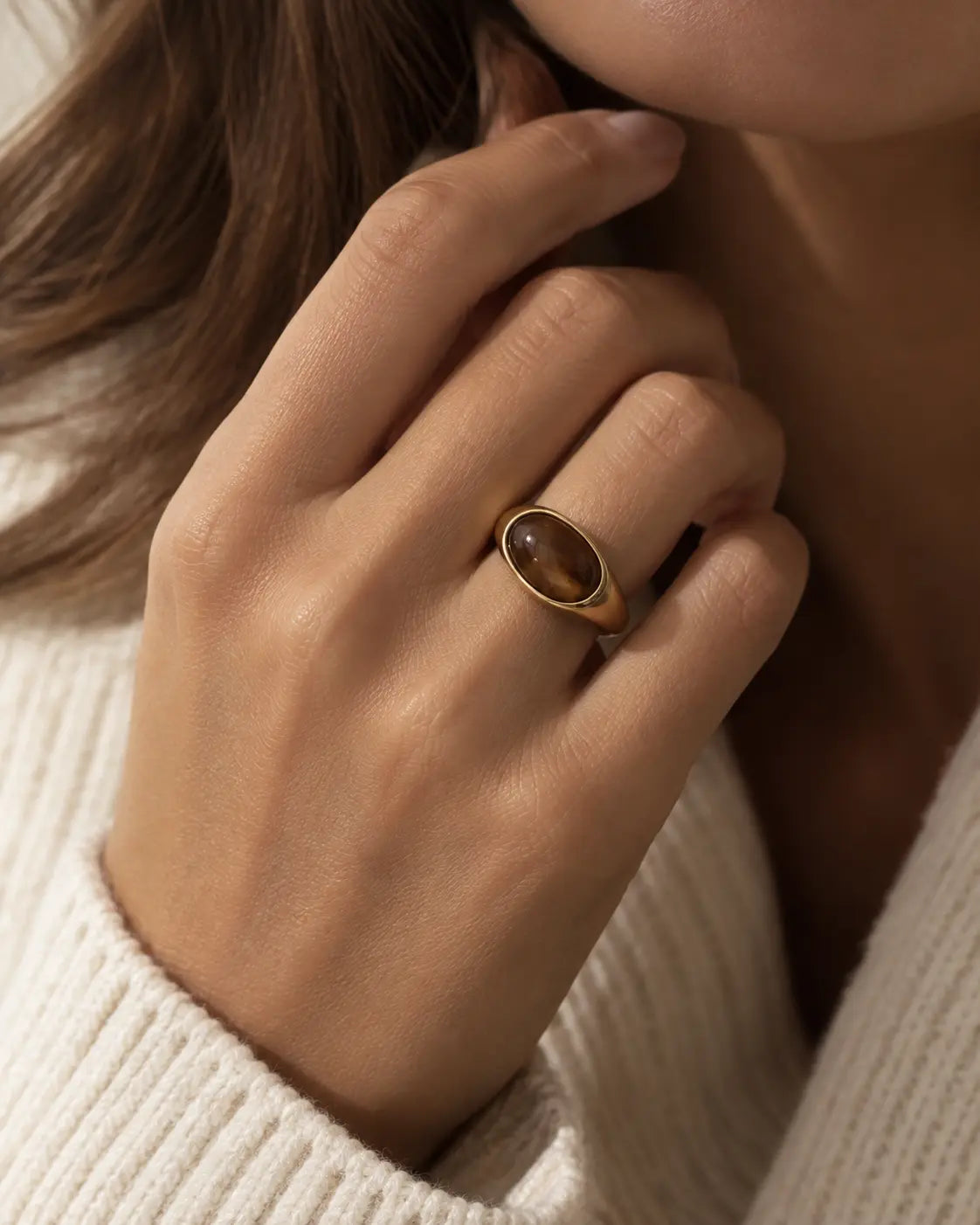 Gold-tone ring with a horizontal brown oval accent, worn on a hand near the chin.