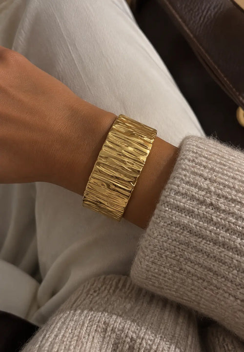 Wide gold-tone cuff bangle with a bark-like texture, close-up on the wrist beside cream knitwear.