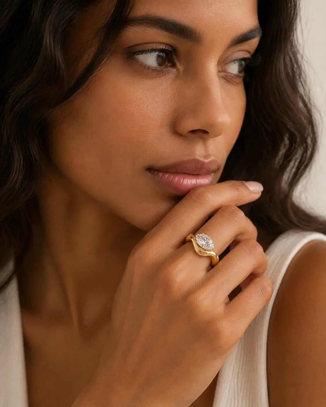 Gold-tone wave band ring with a raised clear stone accent, worn on a hand near the chin.
