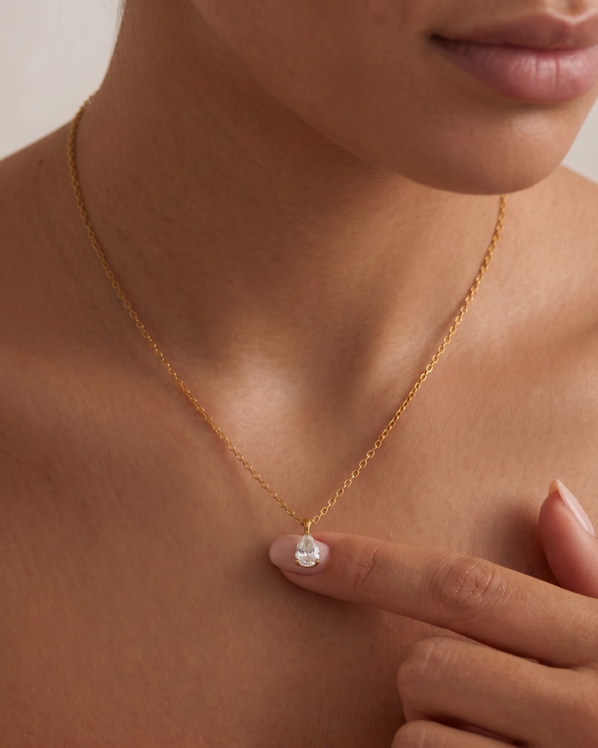 Gold-tone teardrop pendant with a clear stone accent on a fine chain, worn with fingers touching the pendant.