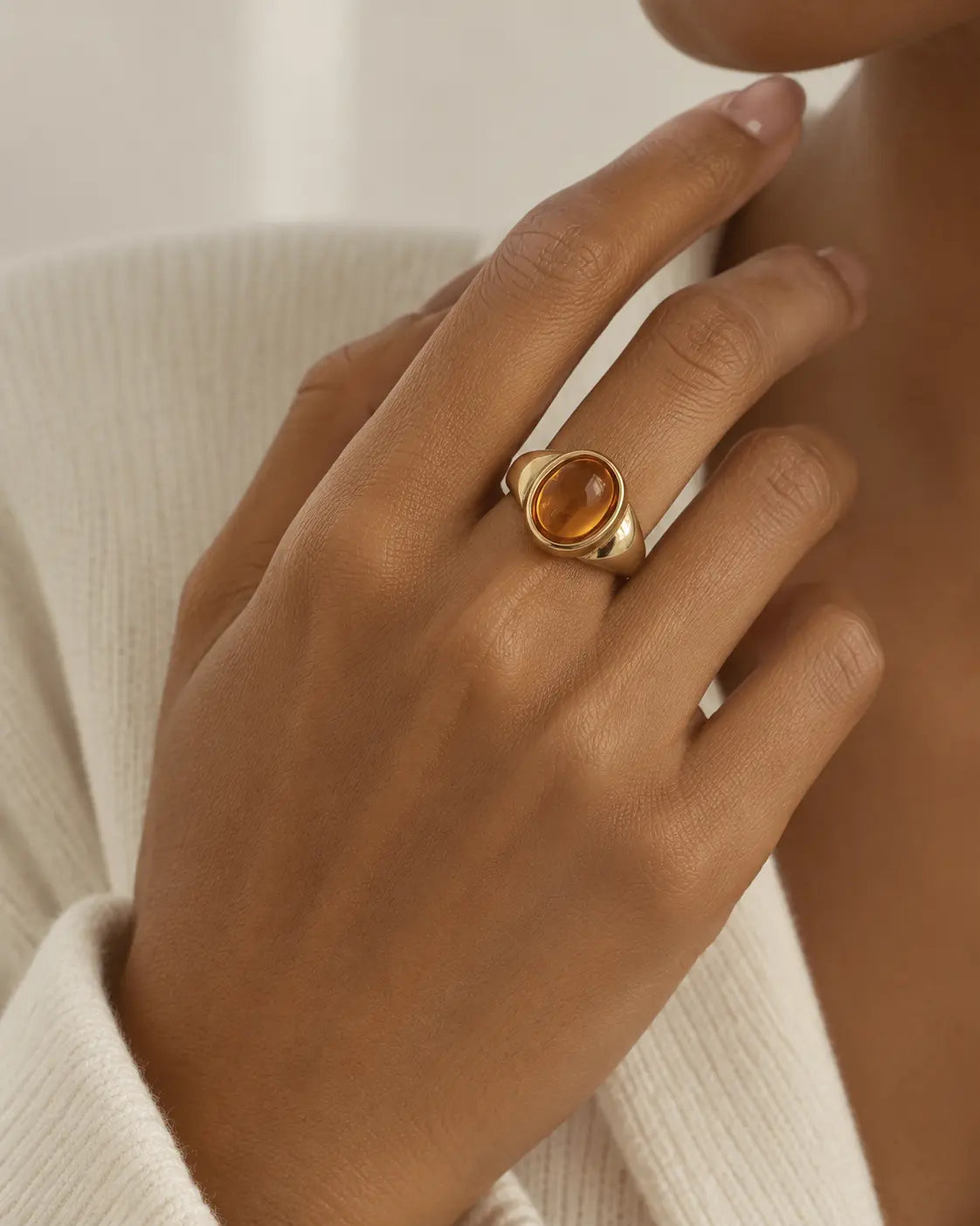 Gold-tone signet ring with a raised warm-brown oval accent, worn on a hand near the chin.