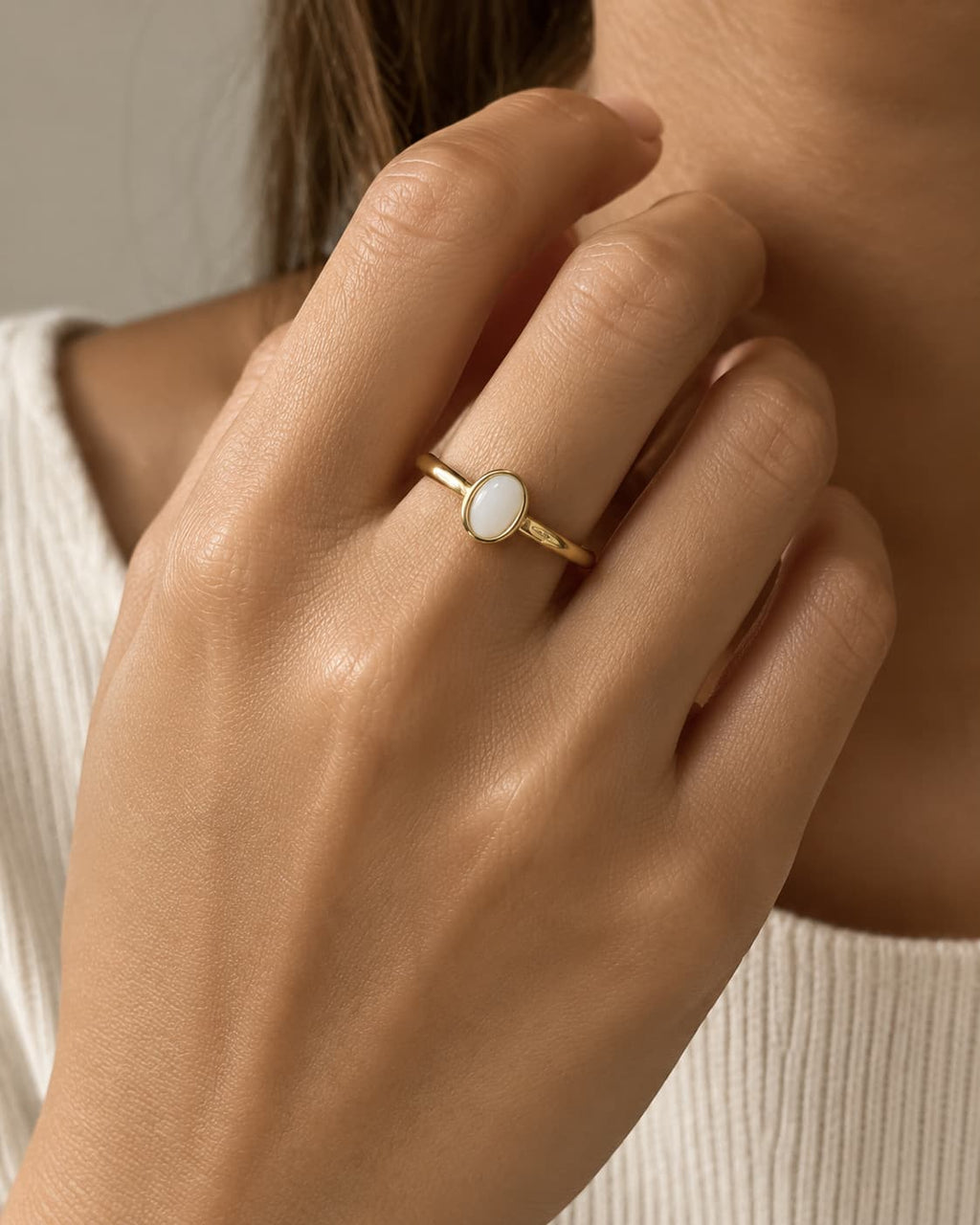 Slim gold-tone ring with a white oval bezel accent, worn on a raised hand.