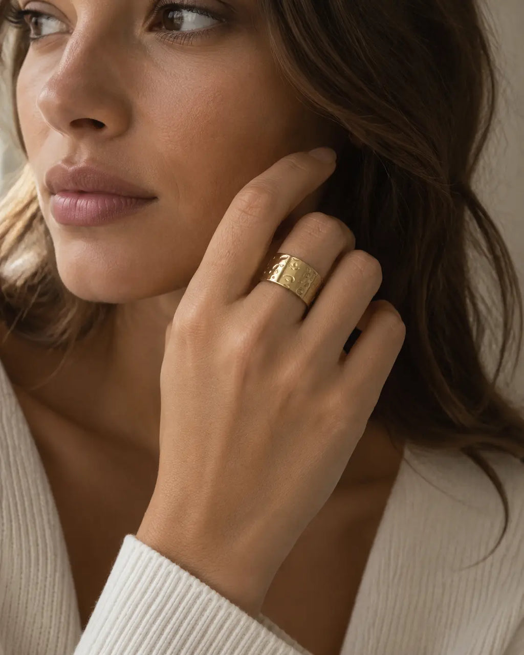 Wide gold-tone ring with a hammered surface, worn on a hand beside the cheek.