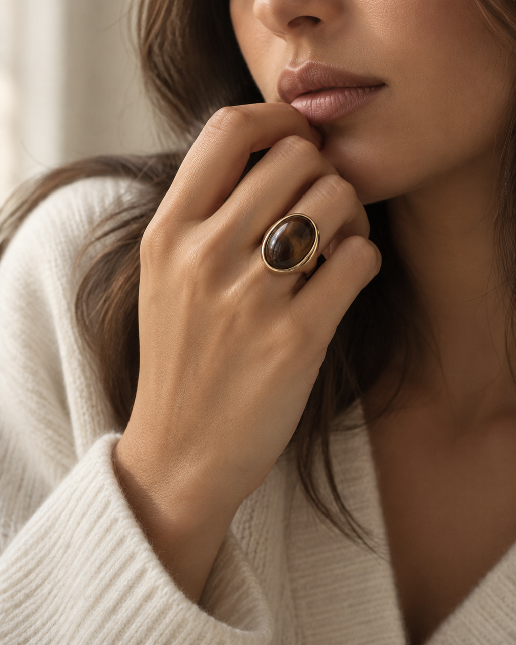 Gold-tone signet ring with a tall dark-brown oval accent, worn on a hand near the chin.