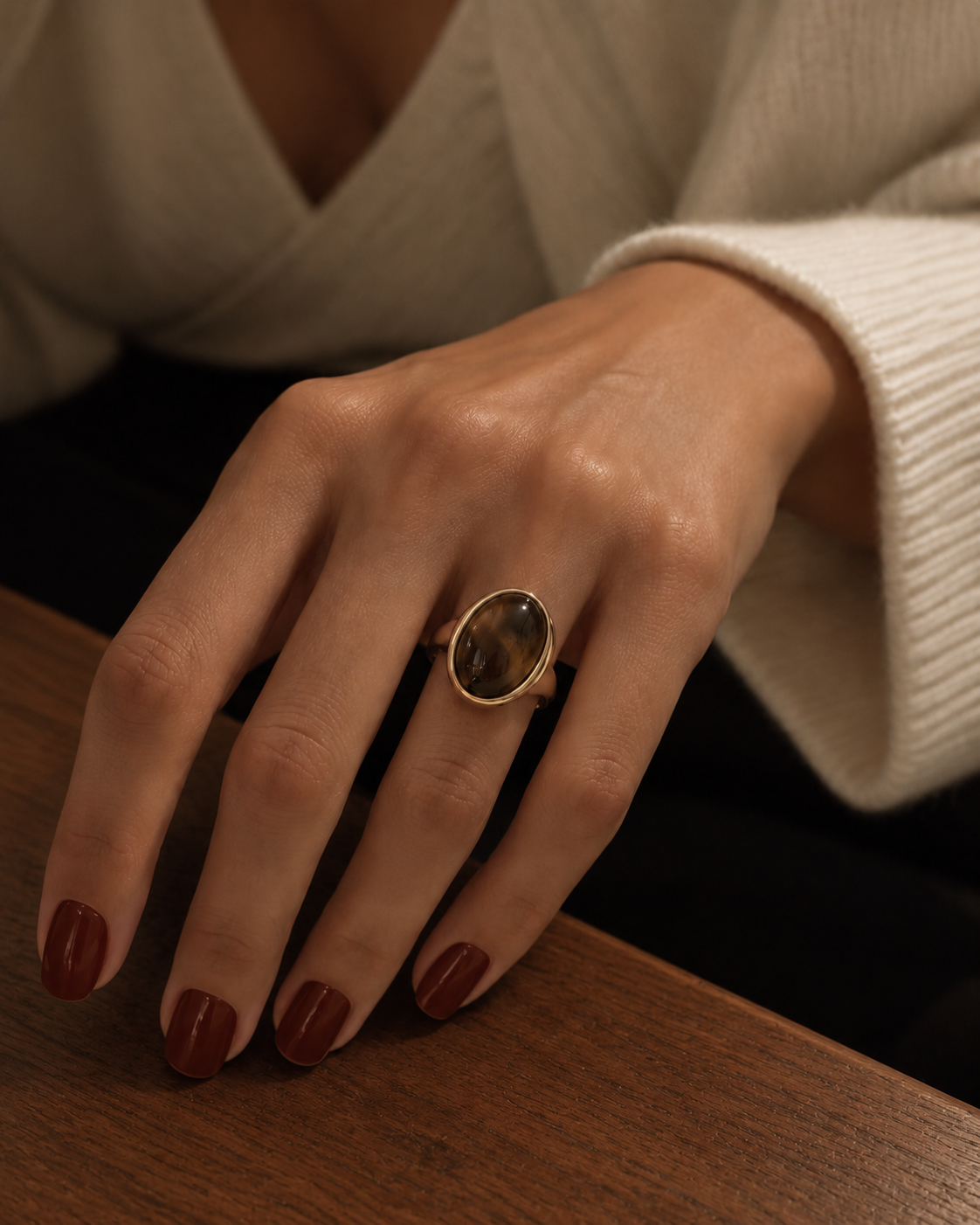 Gold-tone signet ring with a tall dark-brown oval accent, worn on a hand resting beside a wooden surface.