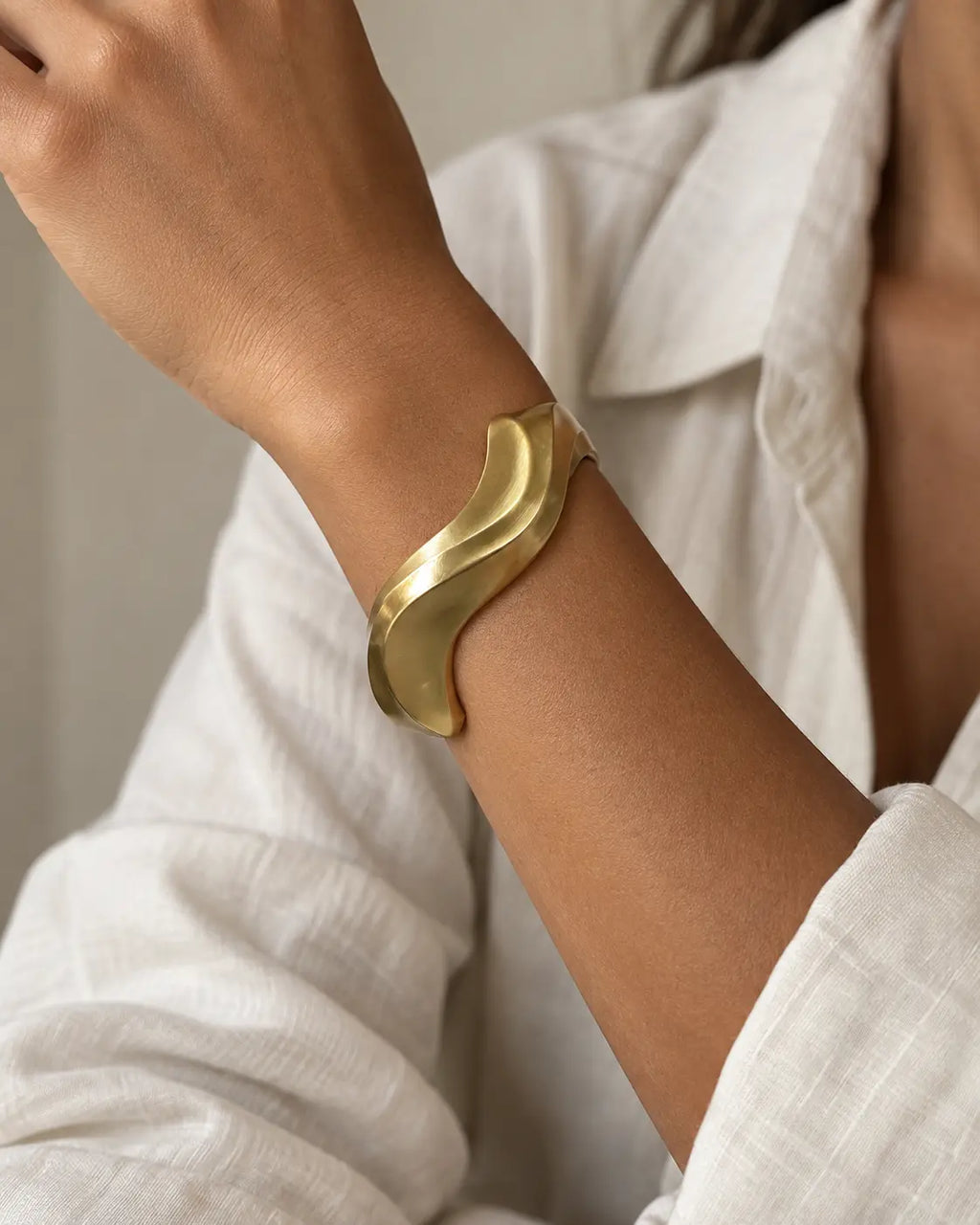 Gold-tone sculptural bangle with an undulating outline, worn on a raised wrist beside white fabric.