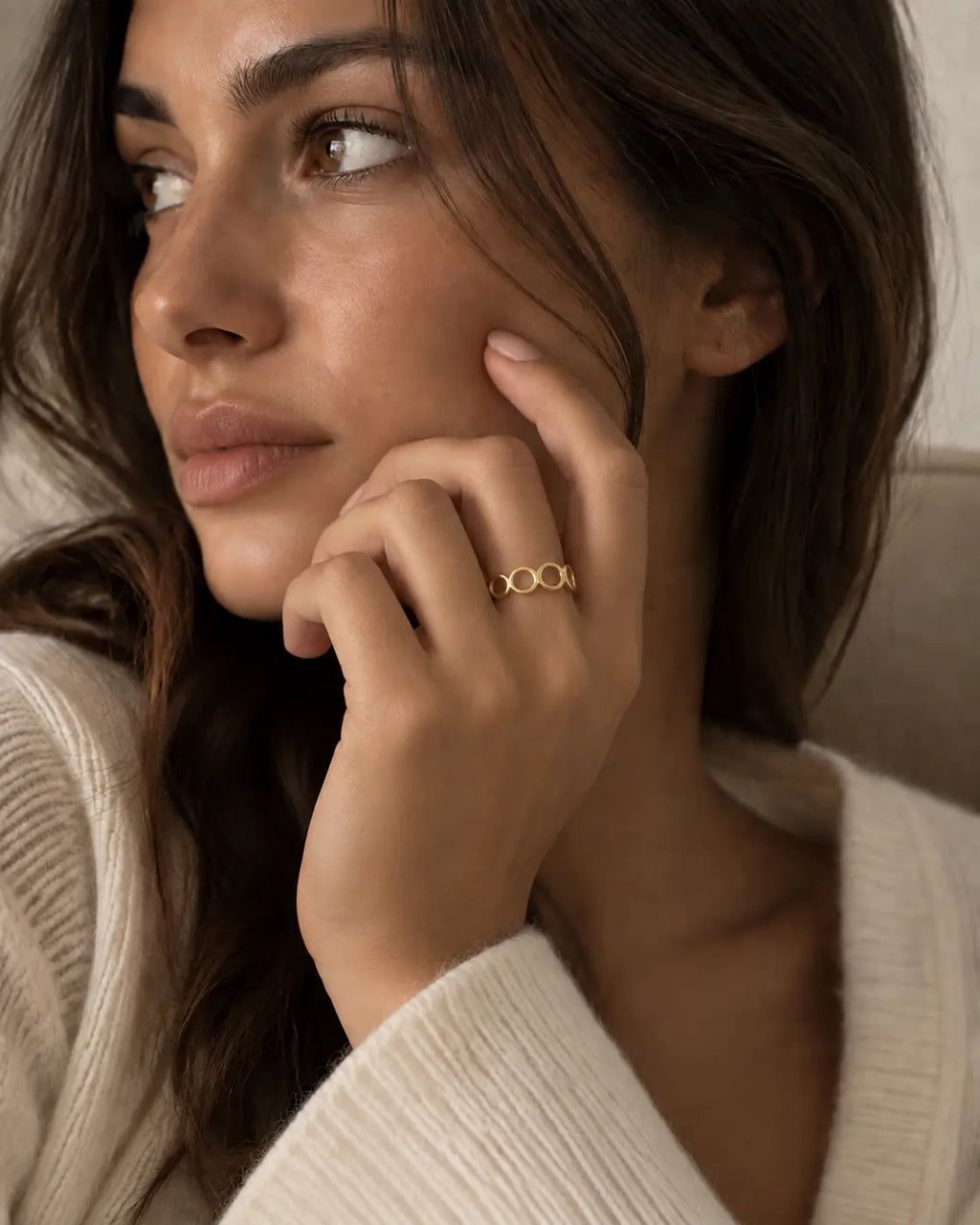 Gold-tone open band ring with linked geometric loops, worn with a hand beside the cheek.